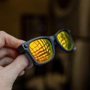 May include: Black sunglasses with yellow-tinted lenses. The lenses reflect a grid pattern, and the frames are a matte black. The sunglasses are held in a hand, with the focus on the eyewear.