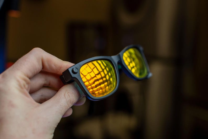 May include: Black sunglasses with yellow-tinted lenses. The lenses reflect a grid pattern, and the frames are a matte black. The sunglasses are held in a hand, with the focus on the eyewear.