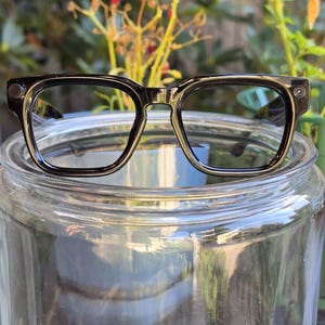 May include: Black-framed eyeglasses with a thick, rectangular silhouette and rounded corners. The frames feature a glossy finish and small, circular metallic accents on the outer top corners of each lens rim, providing a structured look.