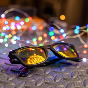 May include: Black-framed glasses with yellow-tinted lenses rest on a textured surface, reflecting a warm, yellow light. A string of colorful LED lights in various colors, including blue, green, and red, is in the background, creating a festive atmosphere.