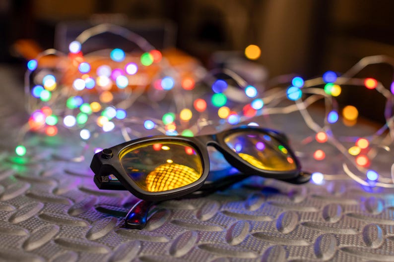 May include: Black-framed glasses with yellow-tinted lenses rest on a textured surface, reflecting a warm, yellow light. A string of colorful LED lights in various colors, including blue, green, and red, is in the background, creating a festive atmosphere.