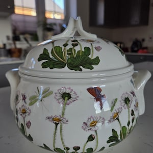 May include: White ceramic serving dish with lid, decorated with a floral pattern of daisies, butterflies, and dragonflies. The dish has two handles and a decorative handle on the lid. The floral design includes green leaves and stems, with yellow and purple accents.