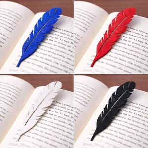 Peut inclure: Quatre marque-pages en forme de plumes, bleus, rouges, blancs et noirs, chacun placé dans un livre ouvert. Les marque-pages sont conçus pour ressembler à des plumes, avec des bords détaillés et un aspect texturé. Les pages du livre ont du texte imprimé.
