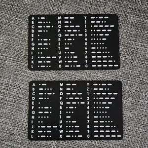Può includere: Due carte nere con simboli Morse bianchi per l'alfabeto e i numeri. Le carte sono rettangolari e hanno una finitura opaca. Sono appoggiate su una superficie grigia strutturata.
