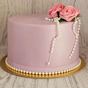 Può includere: Una torta rotonda color lavanda decorata con rose rosa e una collana di perle. La torta è su un vassoio dorato, con un bordo di perle attorno alla base. È un dolce.