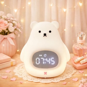 Può includere: Una sveglia bianca a forma di orso con display digitale che mostra le 07:45 AM. L'orologio ha occhi neri e una bocca a forma di piccolo fiocco. Si trova su una tovaglietta di pizzo bianco, con cuori rosa e un vaso di rose rosa sullo sfondo.