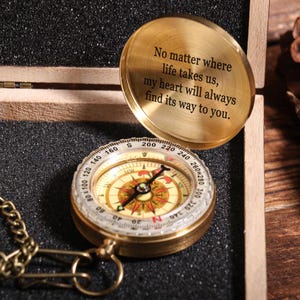 Peut inclure: Une boussole en laiton avec une chaîne, dans une boîte en bois. Le cadran de la boussole comporte des indications directionnelles. Le couvercle de la boîte est ouvert, révélant l'inscription : "No matter where life takes us, my heart will always find its way to you."