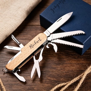 May include: A multi-tool pocket knife with a wooden handle engraved with the name "Michael". The knife features various tools, including a blade, saw, and scissors. A navy blue gift box and a burlap bag are in the background, along with a length of rope, on a dark wood surface.