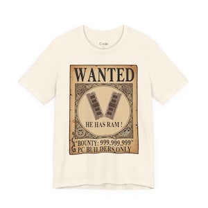 Könnte beinhalten: Cremefarbenes T-Shirt mit einem "WANTED"-Poster-Design. Das Poster hat einen braunen Hintergrund mit einem kreisförmigen Emblem mit zwei RAM-Riegeln und dem Text "HE HAS RAM!" und "BOUNTY: 999.999.999 PC BUILDERS ONLY."