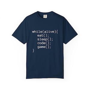 Op de afbeelding: Marineblauw T-shirt met witte tekst: "while(alive){ eat(); sleep(); code(); game(); }." Het T-shirt heeft een ronde hals en korte mouwen.