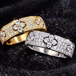 Peut inclure: Deux bagues en diamant. L'une est en or, avec un motif floral et de petits diamants. L'autre est en argent, avec un design floral similaire et de petits diamants. Les deux bagues sont sur une surface noire.