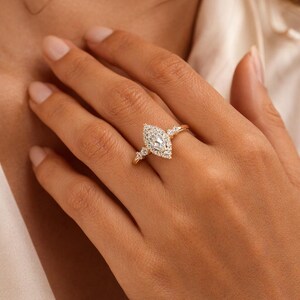Peut inclure: Une bague en or avec un diamant central de taille marquise, entouré d'un halo de petits diamants. La bague comporte des accents de diamants supplémentaires sur la bande. La bague est sur le doigt d'une personne.