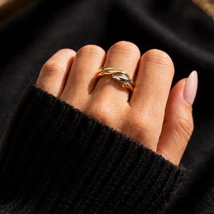 Puede incluir: Un anillo dorado y plateado con un diseño entrelazado. El anillo se lleva en un dedo, sobre un fondo oscuro. El diseño del anillo presenta dos bandas que se cruzan, creando un aspecto único y elegante.