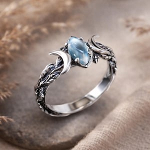 Op de afbeelding: Een zilveren ring met een lichtblauwe ovale edelsteen, geflankeerd door halve manen en bladeren. De ring heeft een gedetailleerde, getextureerde band met een op de natuur geïnspireerd ontwerp. De edelsteen is het middelpunt.