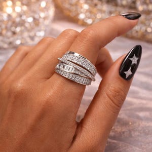 Peut inclure: Une bague en argent avec plusieurs rangées, incrustée de petits pierres précieuses transparentes. La bague présente un design superposé avec des pierres rondes et baguette, créant un effet scintillant. La bague est portée sur un doigt, mettant en valeur ses détails.