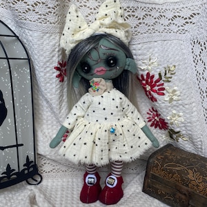 May include: A handmade doll with a green face, gray hair, and a cream-colored dress adorned with black stars. The doll wears a matching bow, red shoes, and has a floral embellishment on the collar. A decorative birdcage and a wooden box are in the background.