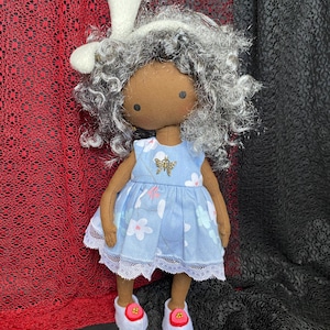 Könnte beinhalten: Handgemachte Puppe mit grauem Lockenhaar, braunem Gesicht und blauem Blumenkleid. Die Puppe trägt ein weißes Stirnband mit Hasenohr und weiße Schuhe mit roten Akzenten. Eine goldfarbene Biene befindet sich auf dem Kleid.