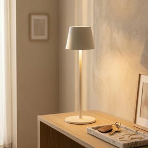 May include: A contemporary cream table lamp with a cylindrical stem and a conical shade. The lamp sits on a wooden table, alongside a marble tray holding keys and jewellery. The background features a neutral-toned wall and a framed picture.