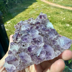 Pode incluir: Um aglomerado de cristais de ametista, mostrando uma gama de tons roxos. Os cristais têm uma textura natural e áspera. Os cristais são segurados em uma mão, com um fundo verde visível.