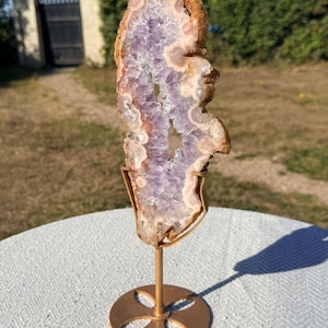 May include: A vertical amethyst geode slice with a jagged, crystalline purple centre and a rough, tan-coloured outer edge. The mineral specimen is securely held upright by a gold-toned metal stand featuring a circular base with four cutouts.