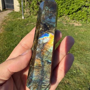 以下が含まれることがあります： 青、金、緑色の虹色の輝きを持つ、研磨されたラブラドライトのクリスタルタワー。 宝石は手で持たれており、その自然な模様と色を示しています。 クリスタルはファセット形状で、先が尖っています。