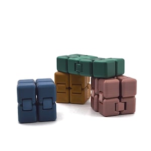 Könnte beinhalten: Eine Sammlung farbiger Fidget-Würfel in Blau-, Braun-, Grün- und Rosatönen. Die Würfel sind in verschiedenen Konfigurationen angeordnet, was ihr ineinandergreifendes Design zeigt. Der Hintergrund ist schlicht weiß.