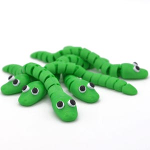 Könnte beinhalten: Mehrere hellgrüne, segmentierte Spielzeugschlangen mit schwarz-weißen Augen liegen auf einer weißen Oberfläche. Die Schlangen bestehen aus flexiblem Material und ihre Körper sind gebogen und beweglich.