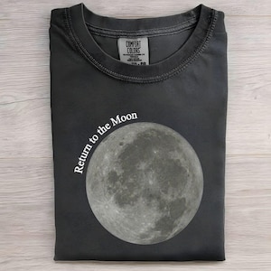 Puede incluir: Camiseta gris oscuro con un gráfico redondo de la luna y el texto "Return to the Moon" en blanco. La camiseta está doblada y tiene una etiqueta en la parte superior central. La tela es suave.