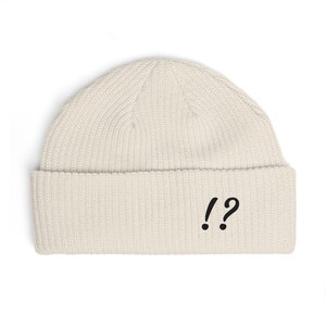 Puede incluir: Gorro de color crema con borde doblado. El gorro presenta un signo de exclamación y un signo de interrogación negros en la parte delantera. El gorro está hecho de un material de punto acanalado.