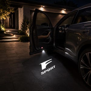 Puede incluir: Una puerta de coche está abierta por la noche, proyectando un logotipo F SPORT blanco brillante en el suelo. El vehículo tiene un exterior oscuro y el panel interior de la puerta muestra detalles en color canela. La luz ilumina el pavimento.