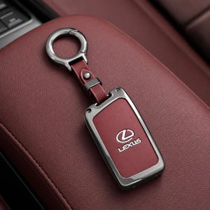 May include: A dark red leather key fob cover featuring a metallic gunmetal frame and a circular ring attachment. The center of the cover displays the silver Lexus logo and the word Lexus in a clean, sans-serif font, matching the key fob's design.