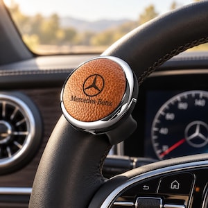 Op de afbeelding: Een cirkelvormig stuurwielaccessoire met een textuurrijk bruin lederen middenstuk met het zwarte Mercedes-Benz logo en tekst. Het embleem is gevat in een gepolijst chromen frame en is gemonteerd op het zwarte lederen stuurwiel van een voertuig.