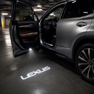 Puede incluir: Un SUV Lexus plateado con la puerta del conductor abierta, que cuenta con una luz de cortesía que proyecta el logotipo blanco de Lexus en el suelo. El interior del vehículo muestra detalles en cuero marrón en la puerta, volante y salpicadero.