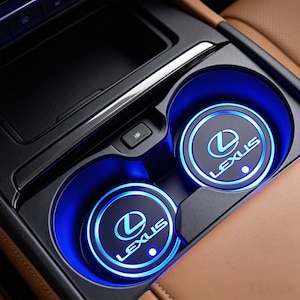 Könnte beinhalten: Zwei kreisförmige, schwarze Auto-Getränkehalter-Einsätze mit dem weißen Lexus-Logo und Schriftzug, beleuchtet durch einen hellblauen LED-Ring. Die Einsätze befinden sich in einem dunkelgrauen Mittelkonsolenfach neben einer beigen Lederpolsterung.