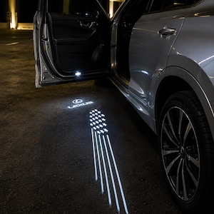 May include: A silver car with an open door projecting a bright white light onto the ground. The projection displays the Lexus logo above a stylized American flag design consisting of stars and vertical stripes, illuminating the dark pavement surface.