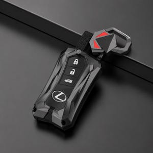 May include: A black Lexus car key fob cover with a geometric, angular design. The case features a central panel with three white icons for locking, unlocking, and boot release, plus the silver Lexus logo. It includes a matching black metal keychain.