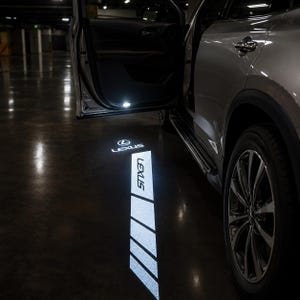 Puede incluir: Un vehículo Lexus plateado con la puerta abierta que proyecta una luz blanca brillante sobre el suelo. La luz muestra el logotipo de Lexus y la palabra LEXUS en un patrón rectangular vertical y segmentado sobre el suelo oscuro.
