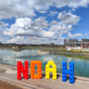 May include: The name "NOAH" spelled out in colorful building blocks. The letters are red, orange, yellow, and blue, arranged on a wooden surface with a lake and cloudy sky in the background. A playful and vibrant display.