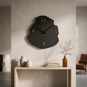 May include: A black wall clock with a unique, organic shape. The clock features gold-colored numerals at the 12, 3, 6, and 9 positions, with matching hands. The clock is mounted on a neutral-colored wall, above a wooden console table.