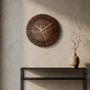 Könnte beinhalten: Eine runde Wanduhr aus Holz mit Sonnenstrahlendesign. Die Uhr hat eine dunkelbraune Oberfläche und weiße Zeiger. Sie ist an einer hellen Wand befestigt. Ein schwarzes Metallregal und eine Vase mit Zweigen befinden sich darunter.