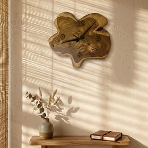 May include: A unique, organic-shaped wall clock made from natural wood. The clock features a dark wood grain pattern and black clock hands. The clock is mounted on a light-colored wall, with a wooden shelf and decorative vase below.