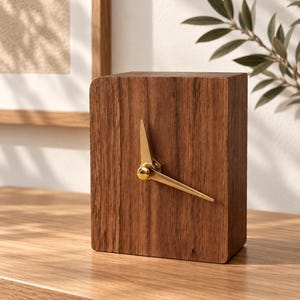 May include: A rectangular wooden desk clock featuring a dark brown wood grain finish and a smooth, flat front surface. The clock is equipped with two slender, polished gold-tone metal hands centred on the face, with no numbers or markings.