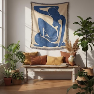 Op de afbeelding: Een geweven sprei in beige en blauw, met een ontwerp van een zittende figuur, hangt boven een houten bank. De bank heeft bruine en gele sierkussens. Potplanten en een vaas met gedroogde grassen maken het interieur compleet.