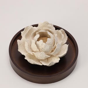 May include: A cream-coloured, paper-like flower sculpture with textured petals, centred on a dark brown, round wooden tray. The flower's centre has a golden hue, and the tray has a smooth, polished finish. The sculpture is a decorative item.
