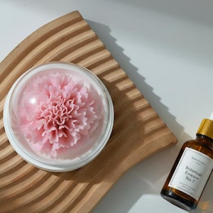 Pode incluir: Uma flor rosa dentro de uma cúpula transparente, sobre uma bandeja de madeira com ranhuras. Ao lado, um frasco de vidro castanho com conta-gotas e tampa dourada, rotulado "Botanical Essence No. 7". Luz suave e natural.