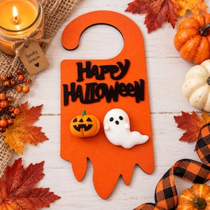 May include: An orange door hanger featuring the black text Happy Halloween, a small orange jack-o'-lantern, and a white ghost figure. The hanger has a curved top for placement on a door handle and a bottom edge shaped like dripping liquid.