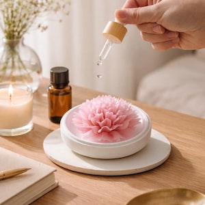 Könnte beinhalten: Ein rosa blütenförmiger Diffusor für ätherische Öle mit einer Pipette, die Flüssigkeit abgibt. Der Diffusor steht auf einer weißen Keramikbasis, neben einer kleinen braunen Flasche und einer brennenden Kerze.