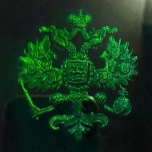 Op de afbeelding: Een levendig groen embleem met een metallic afwerking, voorzien van een tweekoppige adelaar met gespreide vleugels, bekroond door een kroon. De adelaar houdt een scepter in de ene klauw en een rijksappel in de andere, met kleine schilden.