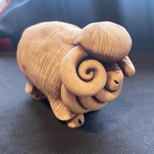 Puede incluir: Una figura de cerámica color canela de un carnero con un patrón de superficie lineal y texturizado en el cuerpo. El carnero presenta cuernos prominentes, grandes y en espiral a ambos lados de la cabeza y una frente redondeada y texturizada.