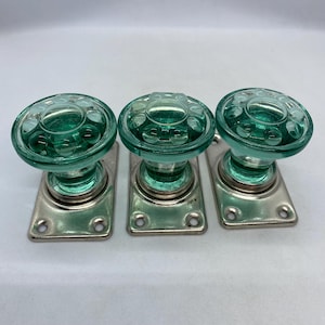 May include: Three round door knobs made of translucent seafoam green glass with a circular embossed pattern on the top surface. Each knob is attached to a rectangular metallic base plate with four screw holes, featuring a polished silver finish.